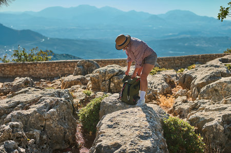 A woman wearing a hat and shorts explores a rocky landscape, looking in her backpack underneath the warm summer sun. The distant mountain range provides a stunning backdrop of natural beauty.の写真素材