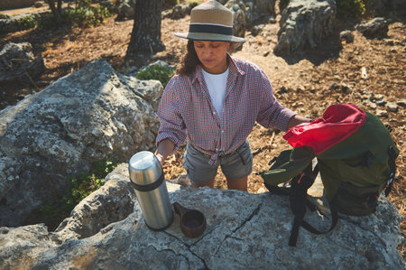 A woman in a hat takes a break during a hike, sipping a drink from a thermos on a rocky trail.の写真素材