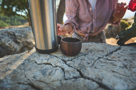 A person in a plaid shirt prepares a hot drink from a thermos on a rocky outdoor surface, embracing nature.の写真素材