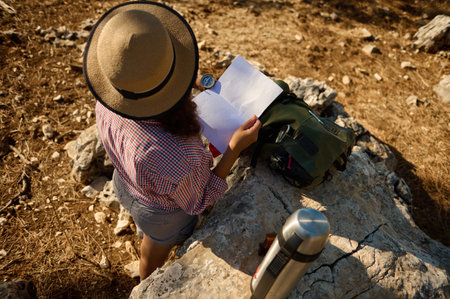 A woman in a hat and checked shirt reading a map while sitting on a rocky terrain. She is surrounded by a travel backpack and a thermos, enjoying the sunny outdoors.の写真素材