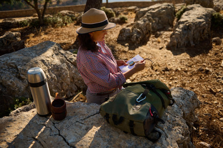 A woman wearing a hat reads a map while sitting on rocks in a sunny, natural setting. A backpack and...