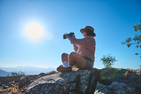 A woman sits on rocky terrain, using binoculars under a clear blue sky. The scene reflects...