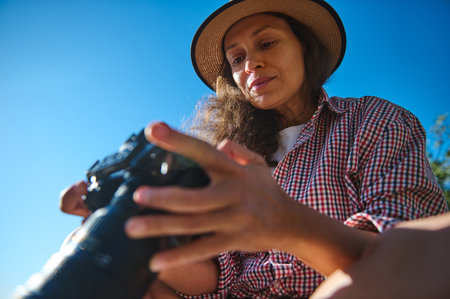 A woman wearing a straw hat and a plaid shirt examines her camera outdoors. The blue sky and natural light create a vibrant setting, symbolizing adventure and creativity.の写真素材