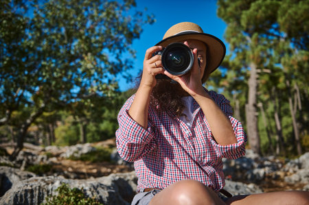 A woman wearing a hat holds a camera, capturing moments in a lush forest. The bright day highlights her passion for photography amidst nature. An image of exploration and creativity.の写真素材