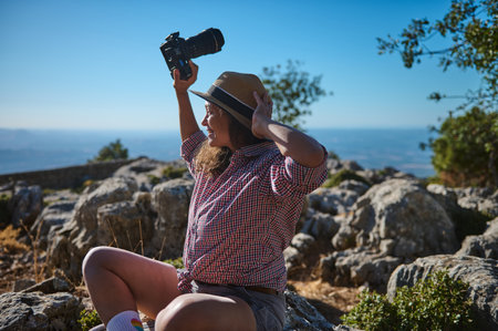 A cheerful photographer raises her camera in excitement while seated in a scenic, rocky landscape...