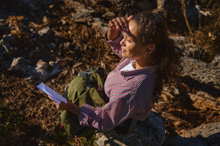 A woman with curly hair sits on rocks in the outdoors, holding a map. Dressed in a checkered shirt, she shades her eyes from the sun, suggesting a hiking or exploring adventure.の写真素材
