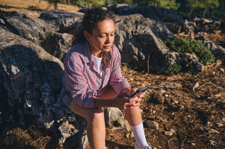 A woman sits on rocky terrain, enjoying a serene outdoor moment while using her smartphone. Sunlight...