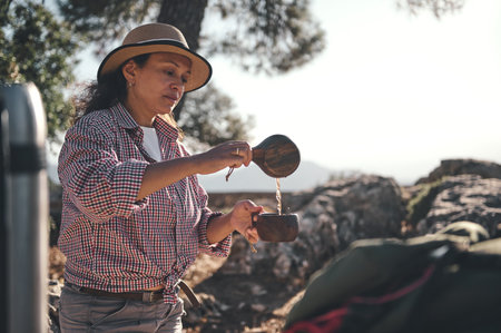A woman in a hat pours coffee outdoors, surrounded by nature. Enjoying a relaxing moment, she is captured in a serene and tranquil setting, showcasing leisure and adventure.の写真素材