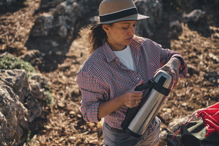 A woman in a checked shirt and hat pours a hot drink from a thermos during a hiking adventure. She seems relaxed and content, embracing the tranquility of nature.の写真素材