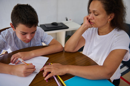 A mother helps her son with his homework at a wooden table. The boy is focused on solving math problems in a notebook while the mother supports him.の写真素材