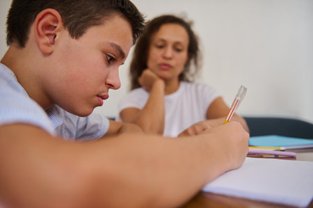 A young student concentrating on writing while a tutor observes attentively. The scene depicts a...