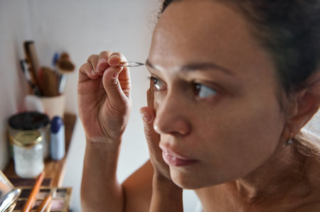 A woman meticulously plucks her eyebrows with tweezers, concentrating on self-care. The scene captures a moment of personal grooming in a bathroom setting.の写真素材