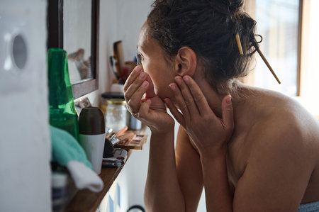 A woman examines her face in the mirror during her morning skincare routine, focusing on self-care and beauty in a cozy setting.の写真素材