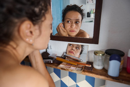 A thoughtful woman looks at her reflection while applying makeup, surrounded by toiletries in the bathroom. The image conveys contemplation, beauty routine, and self-care.の写真素材