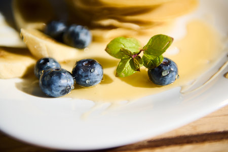 A detailed view of fresh blueberries and mint leaves on pancakes drizzled with syrup. Sunlight enhances the vibrant colors, capturing the freshness and appetizing appeal of this breakfast dish.の写真素材
