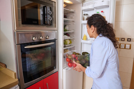 A woman is seen arranging fresh fruits and vegetables in a refrigerator. The modern kitchen setting features a microwave and oven. This image conveys themes of healthy lifestyle and organization.の写真素材