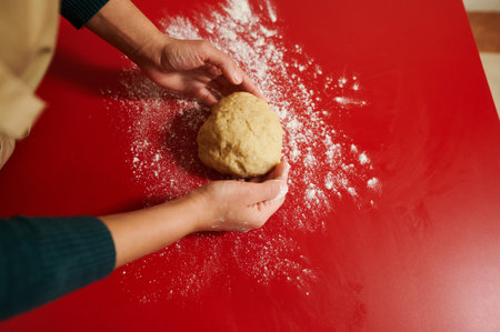 Two hands skillfully knead a ball of dough on a red countertop dusted with flour, reflecting the art of home baking. The vibrant background adds warmth to this culinary scene.の写真素材