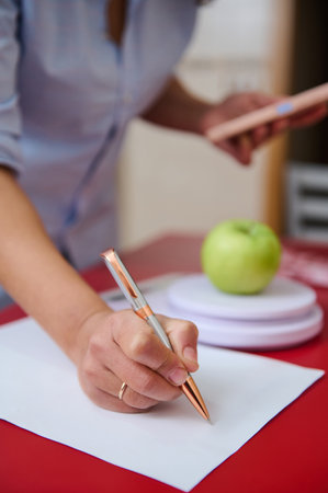 A person takes notes with a pen, while an apple is nearby on a red table. This scene conveys focus,...