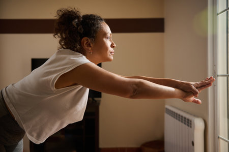 A woman is practicing yoga indoors, stretching forward with focus and calmness. The natural light...