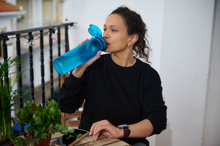A woman sips water from a blue bottle on her balcony, surrounded by potted plants, capturing a...