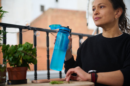 A woman sits on a balcony, holding a blue water bottle, surrounded by plants. She appears relaxed in...