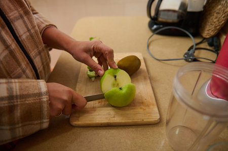 A person carefully slices a fresh green apple on a wooden board in the kitchen. The cozy setting suggests a focus on healthy eating and meal preparation, with a kiwi nearby.の写真素材