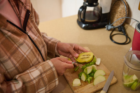 A woman slices kiwi and green apple on a wooden cutting board. A blender, coffee maker, and other kitchen tools are on the counter. The scene evokes a homey, healthy food preparation setting.の写真素材
