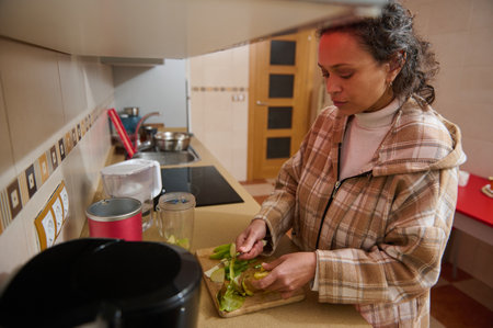 A woman is standing in her kitchen, preparing fresh vegetables on a cutting board. The warm ambiance of the kitchen suggests a cozy and comfortable home environment.の写真素材