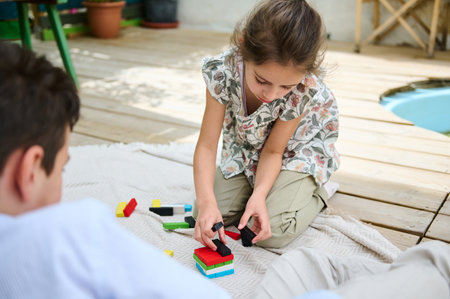 A young girl is focused on building with colorful blocks on an outdoor wooden deck. Her expression reflects concentration and creativity. The setting is casual and relaxed.の写真素材