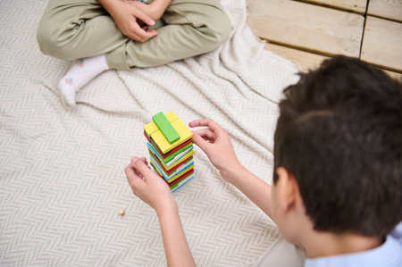 Two children are engaged in a colorful block stacking game, sitting on a soft, patterned blanket. The scene evokes a sense of concentration, fun, and creativity, ideal for family playtime.の写真素材