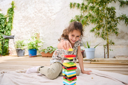 A young girl enjoys building a tower with colorful blocks in a bright outdoor space. Surrounded by greenery and potted plants, she concentrates on creating a balanced structure on a soft blanket.の写真素材