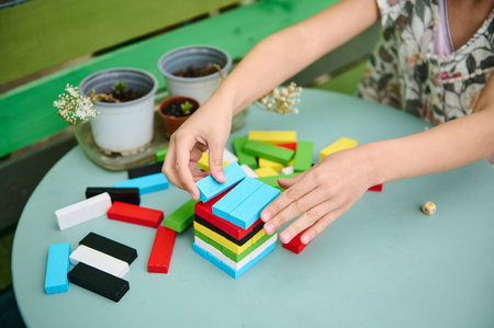 A child is stacking colorful wooden blocks on a green table, surrounded by small potted plants. The...
