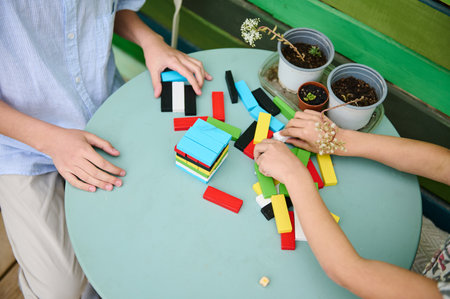 Two children engage in creative play using colorful wooden blocks on a green outdoor table. Nearby,...