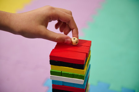A child's hand is carefully adding a small wooden block to a colorful tower, balancing concentration and creativity on a bright, colorful play mat. This image captures the essence of childhood play.の写真素材