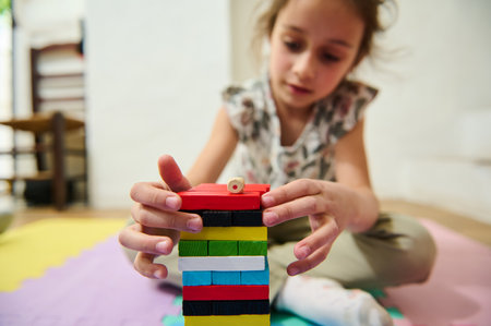 A young girl attentively builds a colorful tower with wooden blocks. Her concentration and creativity are evident as she carefully places each piece. The home setting is cozy and inviting.の写真素材