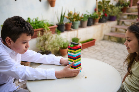 Two children engage in a block stacking game at an outdoor table, concentrating deeply. Surrounded by plants, their expression shows focus and anticipation, creating a warm and playful atmosphere.の写真素材