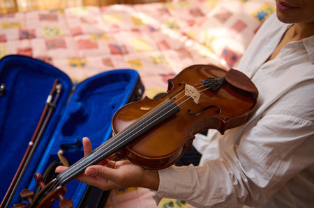 A person in a white shirt carefully holds a violin near a blue case on a colorful, patterned bedspread. The warm tones and soft lighting create a sense of calm and musical appreciation.の写真素材