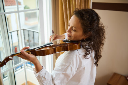 A woman smiles gently as she plays the violin by a sunlit window. The warm ambiance and her focused expression convey a sense of peace and creativity, capturing a moment of musical inspiration.の写真素材