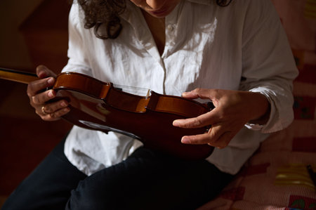 A person in a white shirt sits indoors, closely inspecting a violin under natural light, conveying...