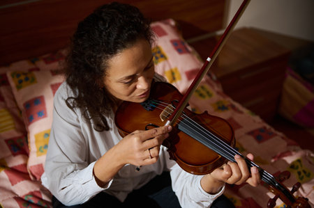 A focused woman plays the violin while sitting on a colorful bedspread in a warmly lit room,...