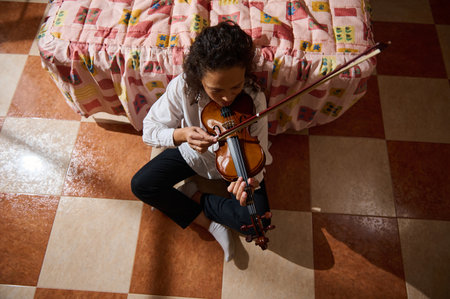 A young girl plays the violin with focus and dedication, seated on a checkered tile floor beside a bed adorned with a colorful quilt, evoking a cozy, creative atmosphere.の写真素材