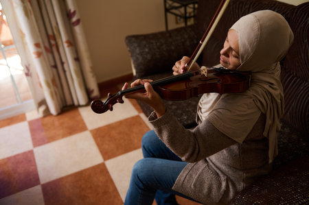 A woman wearing a hijab is seated on a sofa, playing the violin, enveloped in a warm and homey atmosphere.の写真素材