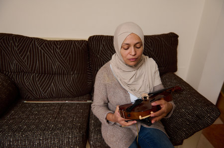 A woman in a beige hijab sits on a patterned sofa, gently holding a violin with closed eyes,...