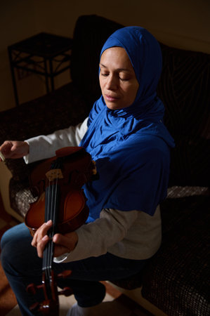 A woman wearing a hijab sits and plays the violin, eyes closed in concentration. The soft lighting...