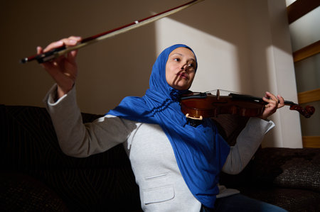 A woman wearing a blue hijab and gray blazer skillfully plays the violin, focusing intently on her...