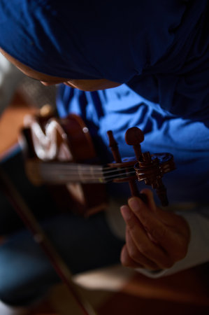 A woman wearing a blue headscarf tunes her violin in soft, warm lighting. Focused on the musician's hands and the instrument, capturing the mood of concentration and precision.の写真素材