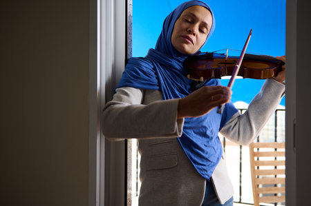 Serene moment as a woman in a headscarf plays the violin with closed eyes, basking in the sunlight on a balcony.の写真素材