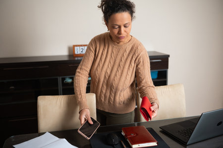 A woman in a cozy home office setting organizes her desk items while holding a smartphone and a red wallet, surrounded by a laptop, notebook, and stack of papers.の写真素材