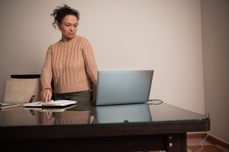 A woman stands at her desk using a laptop, while referencing an open notebook. The setting suggests...