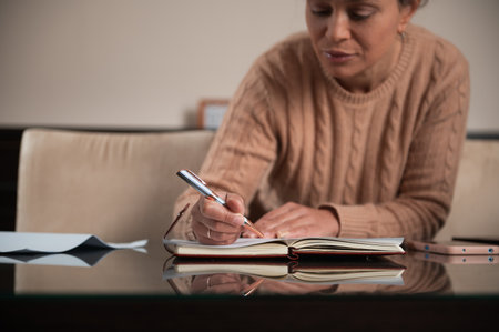 Focused woman writing in a notebook on a reflective table, capturing thoughts.の写真素材